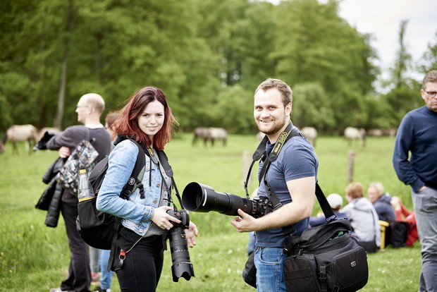 Eine Fotografin und ein Fotograf mit Fotoausrüstung stehen auf einer Wiese, wo im Hintergrund Menschen und Pferde zu sehen sind.