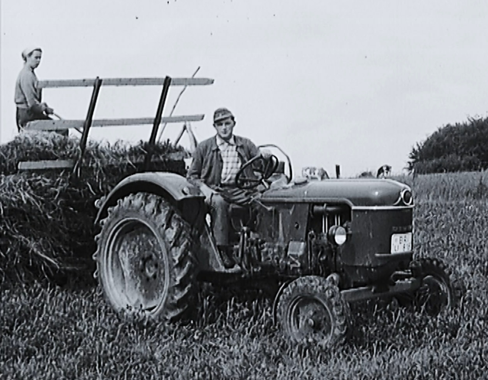 Das Bild zeigt einen Bauern und eine Bäuerin auf einem Traktor mit Anhänger.
