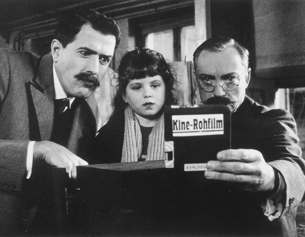 Zwei Männer und ein Mädchen schauen auf eine Schachtel mit der Aufschrift "Kine-Rohfilm".