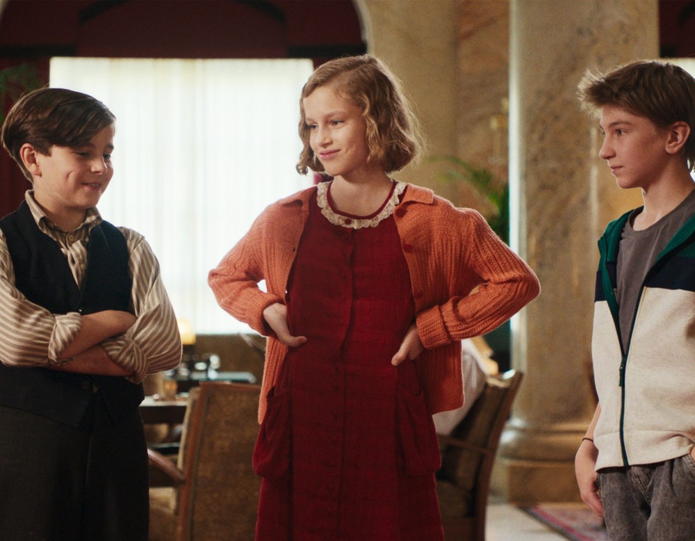 Ein Filmstill aus "Das geheime Stockwerk". Drei Jugendliche stehen nebeneinander - zwei Jungen und ein Mädchen - und lächeln.